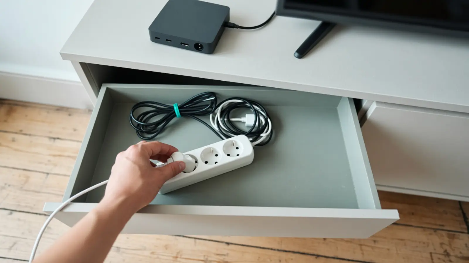 Eine Hand zieht in der offenen Schublade einer Kommode den Stromstecker einer kleinen Streaming-Box ab